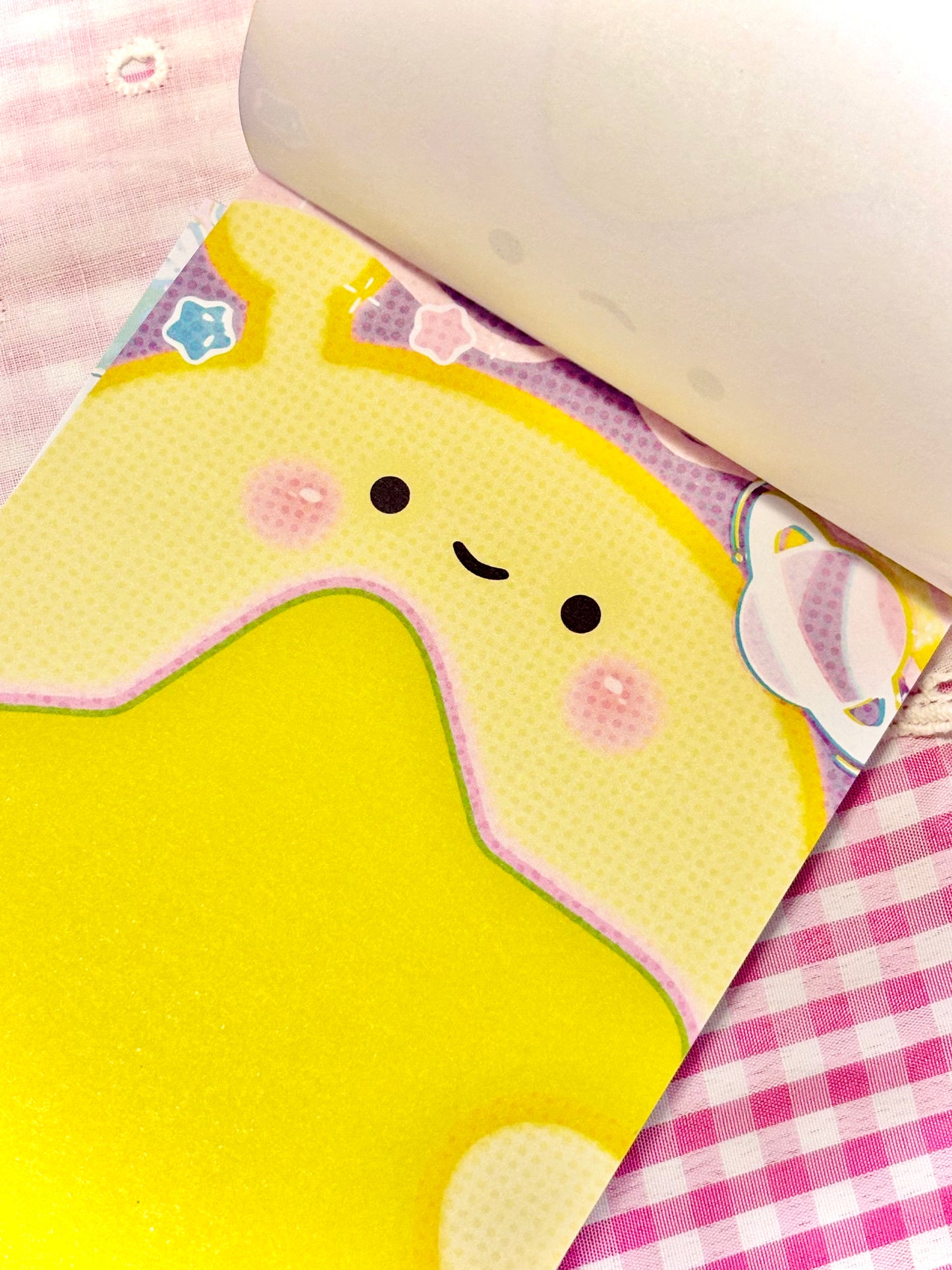 A close-up of a pastel yellow note page featuring a cute cartoon character face, placed on pink-and-white checkered fabric next to a white rolled paper.