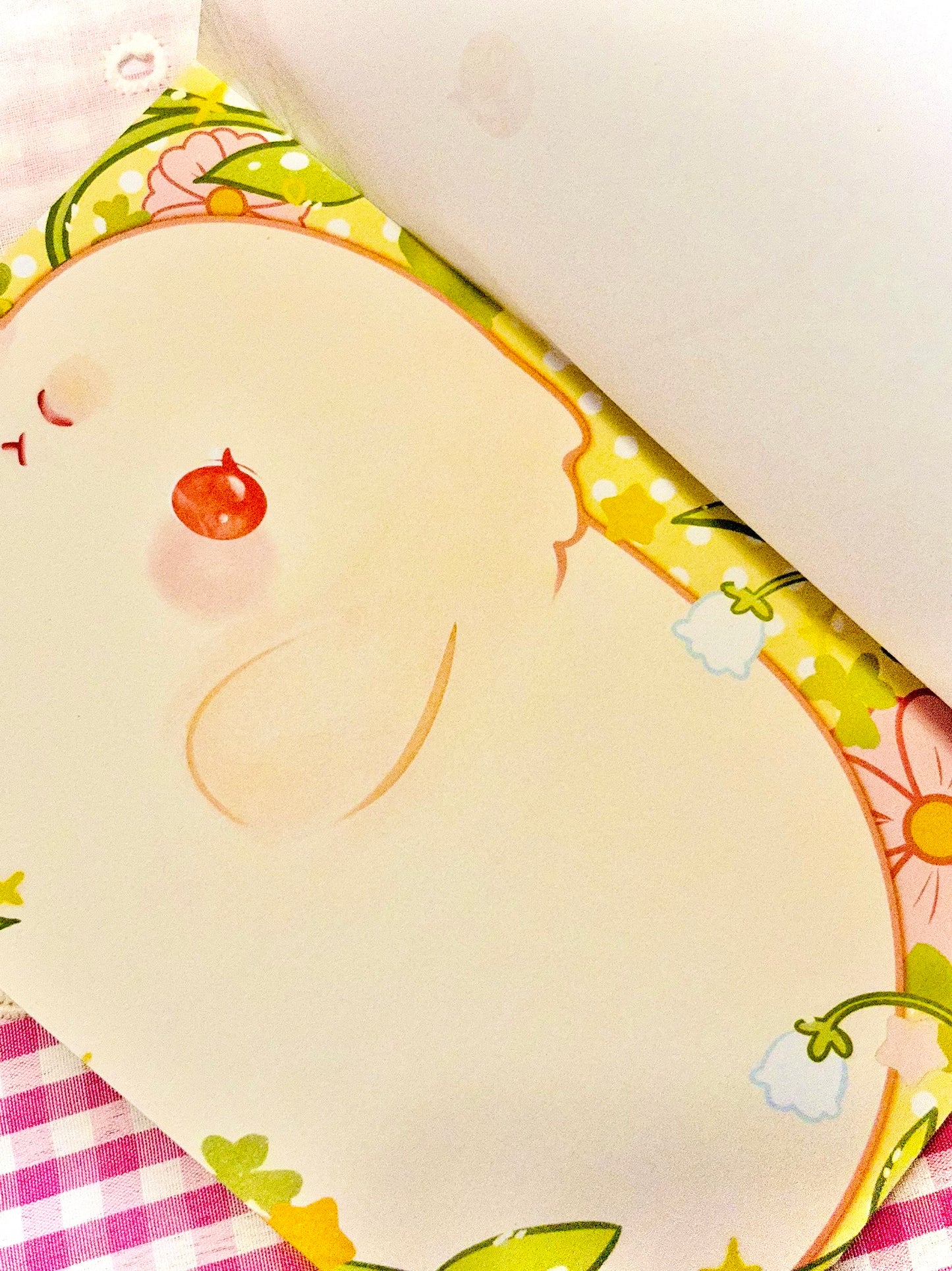 A close-up of a notebook or journal page with a cream-colored cover featuring a featuring a cute white bunny with red eyes and floral border, partially open to reveal blank white pages inside.