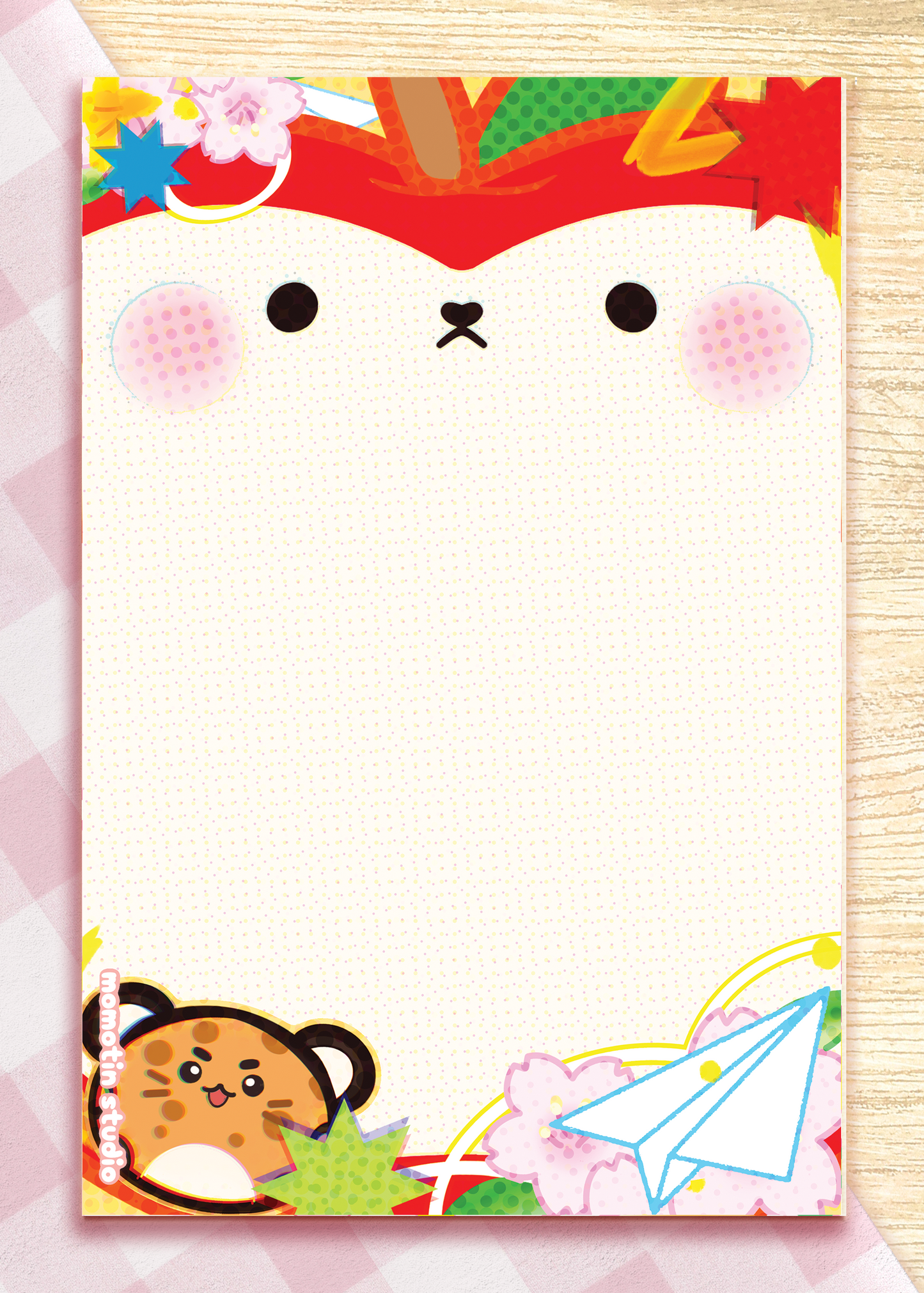 A rectangle notepad with a cute cartoon apple face on the cover, decorated with seasonal motifs including cherry blossoms, a paper airplane, and a cat character at the bottom. The notepad is photographed flat on a light wood surface with a pink patterned sheet partially visible behind it.