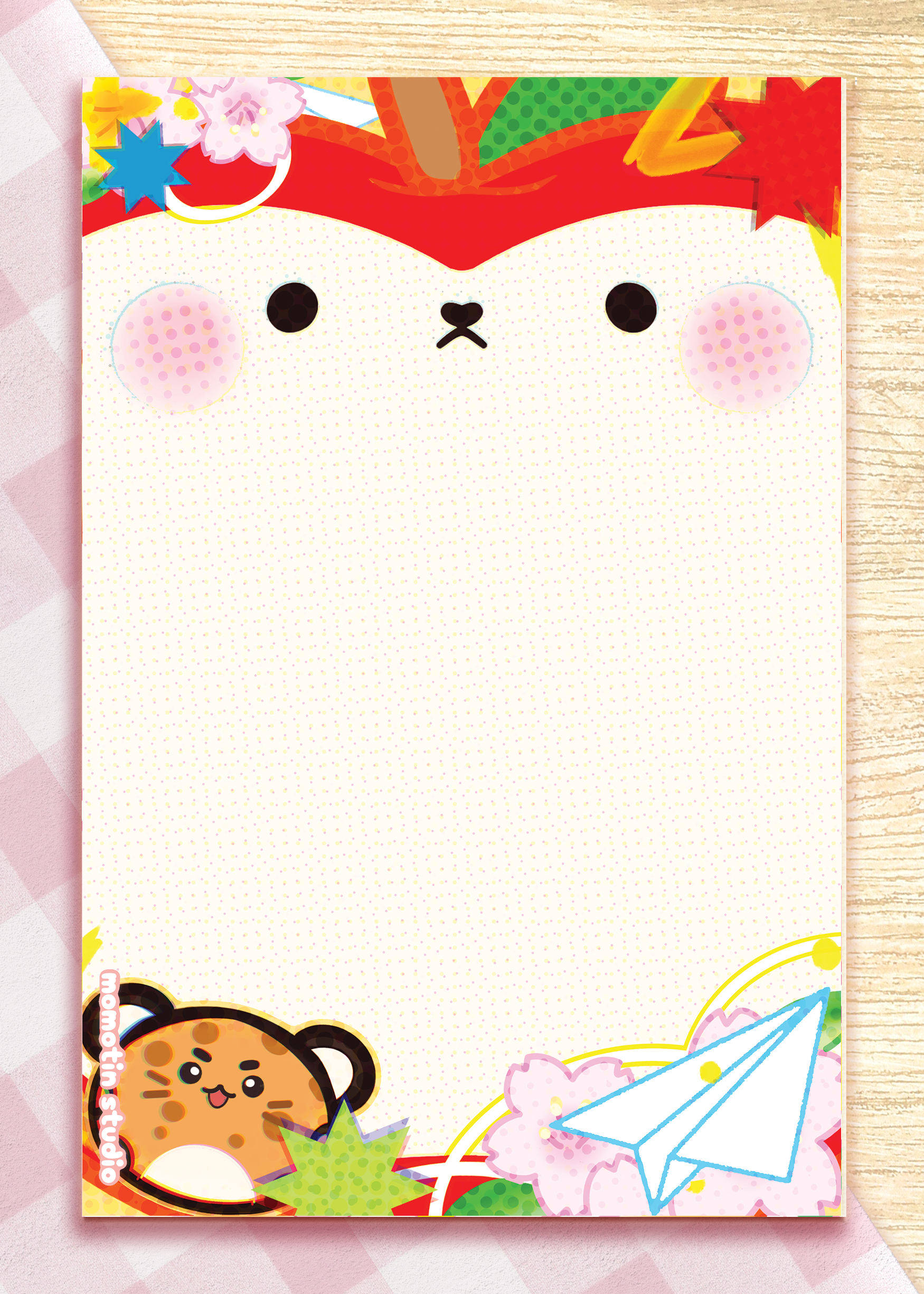 A rectangle notepad with a cute cartoon apple face on the cover, decorated with seasonal motifs including cherry blossoms, a paper airplane, and a cat character at the bottom. The notepad is photographed flat on a light wood surface with a pink patterned sheet partially visible behind it.