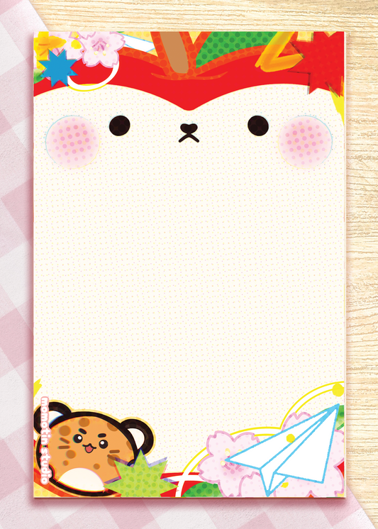 A rectangle notepad with a cute cartoon apple face on the cover, decorated with seasonal motifs including cherry blossoms, a paper airplane, and a cat character at the bottom. The notepad is photographed flat on a light wood surface with a pink patterned sheet partially visible behind it.