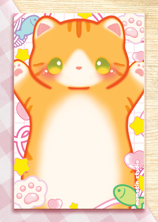 A cute illustrated notepad or card featuring a large orange tabby cat face and paws on a pastel patterned background, photographed flat on a light wood surface.
