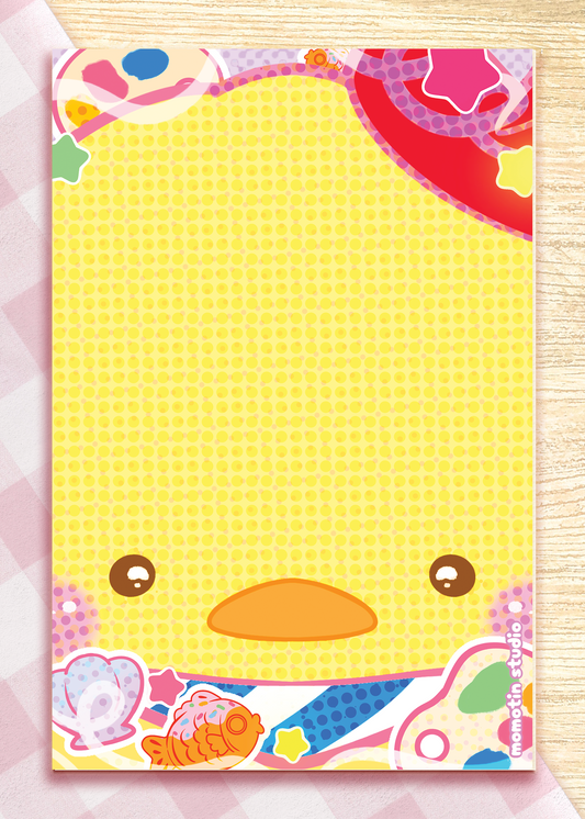 A pastel yellow notepad with a cute cartoon duck face and colorful decorative border, photographed flat on a pink-and-white checkered surface.