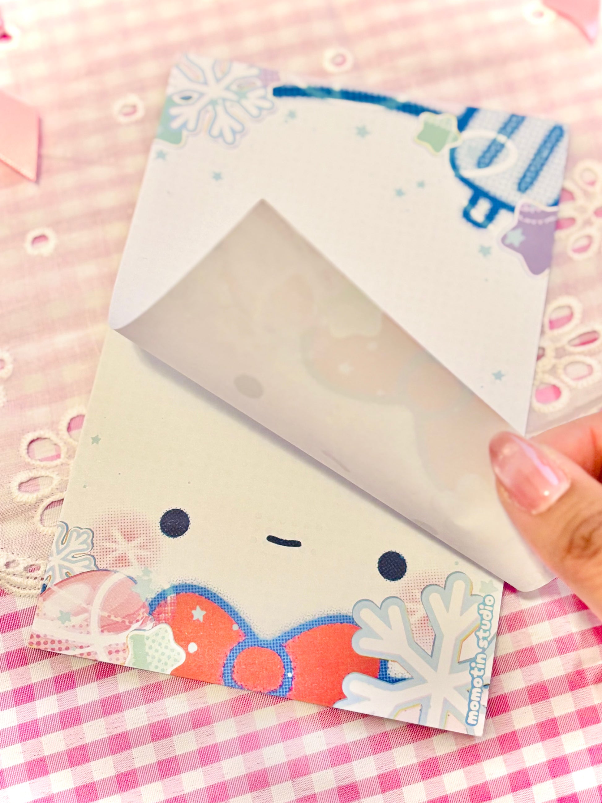 A hand holds open a cute winter-themed notepad. The page features a smiling cartoon face, snowflakes, and a red bow on a pink and white checkered background.