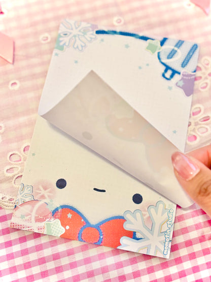 A hand holds open a cute winter-themed notepad. The page features a smiling cartoon face, snowflakes, and a red bow on a pink and white checkered background.