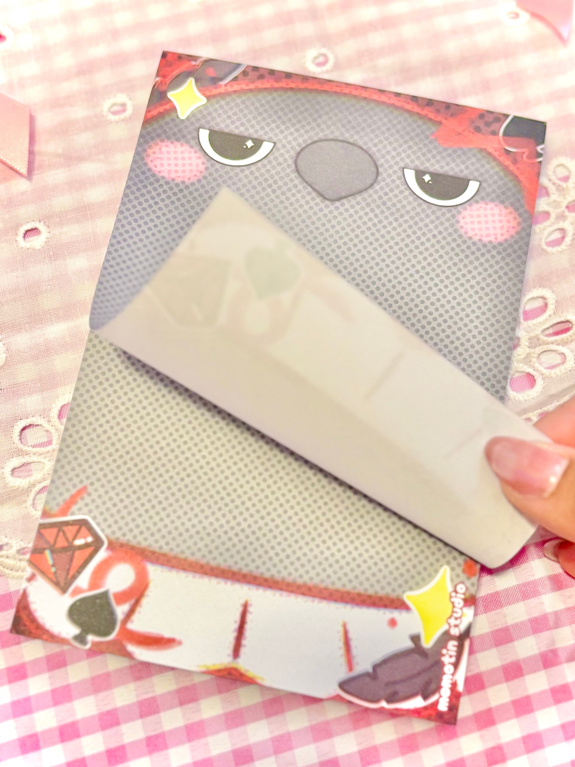 A hand is peeling off a leaflet from a printed notepad featuring a cartoon crow face and black and red graphics, set against a pink and white checkered fabric background.