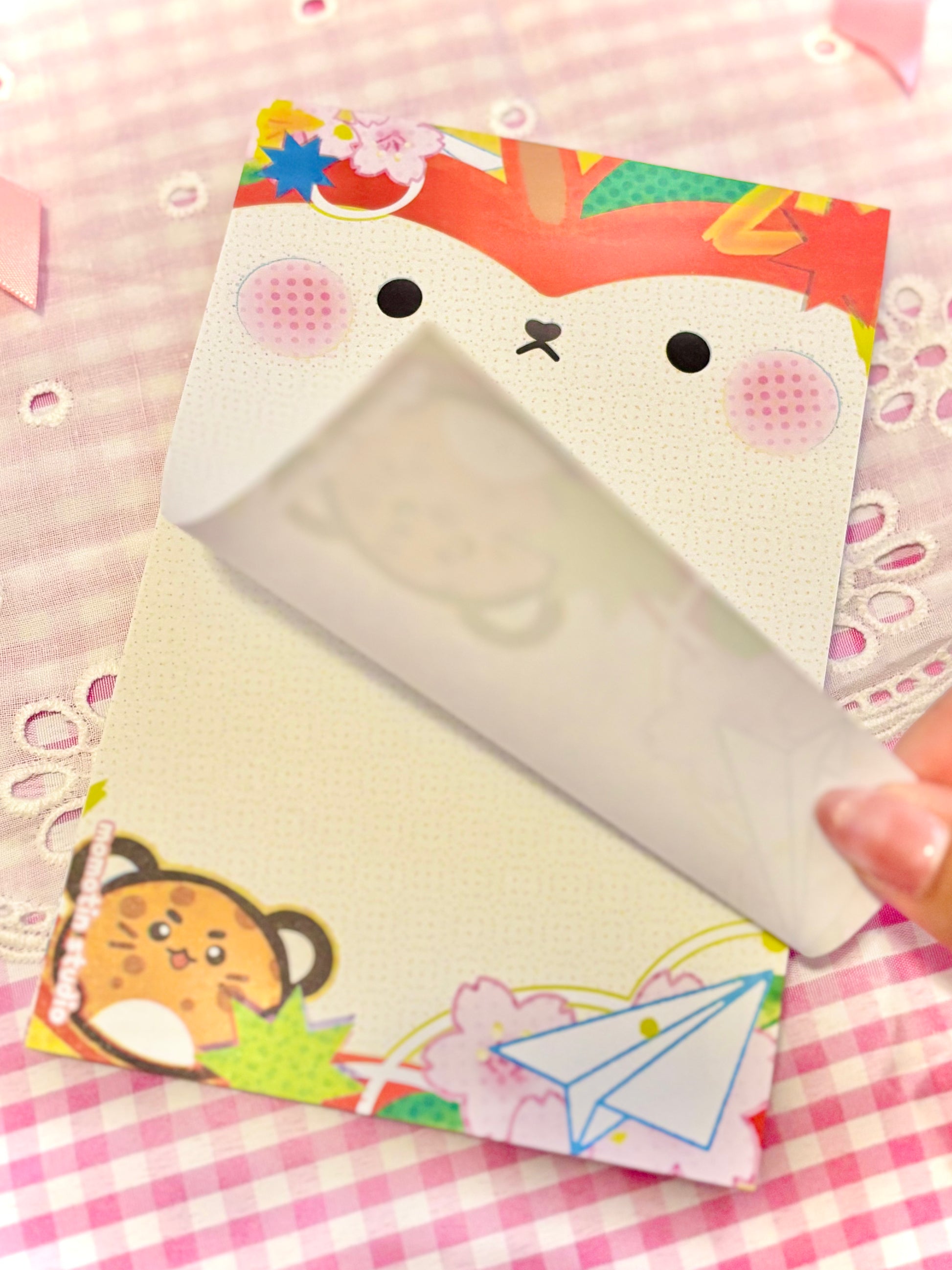 A close-up of a cute stationery notepad with a cartoon apple face on the cover, being opened to reveal a white lined writing surface. A hand is holding the right edge of the notepad.