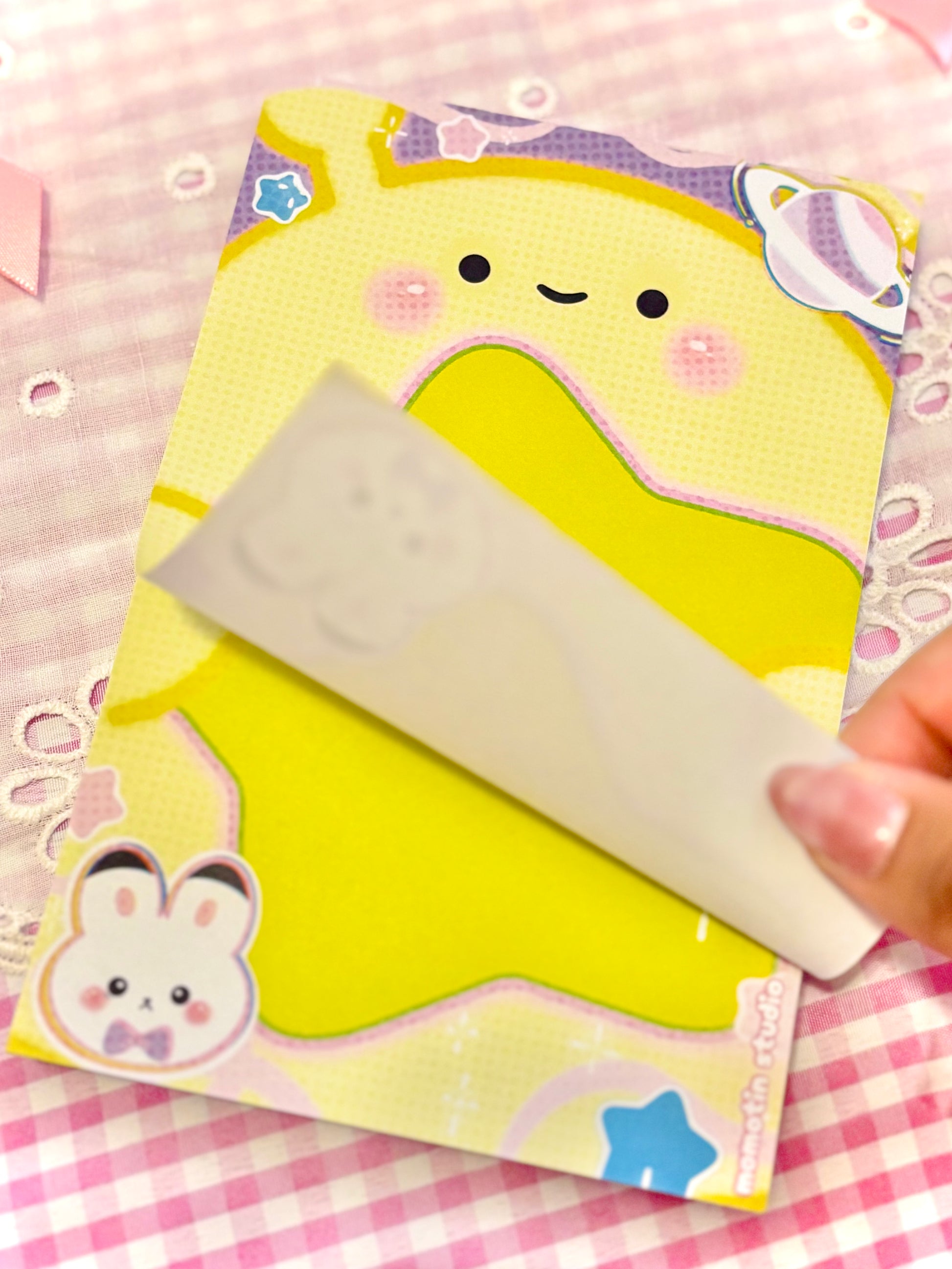 A close-up of a pastel yellow cartoon notepad with a cute bunny and star illustrations, being held by a hand over a pink gingham fabric background.