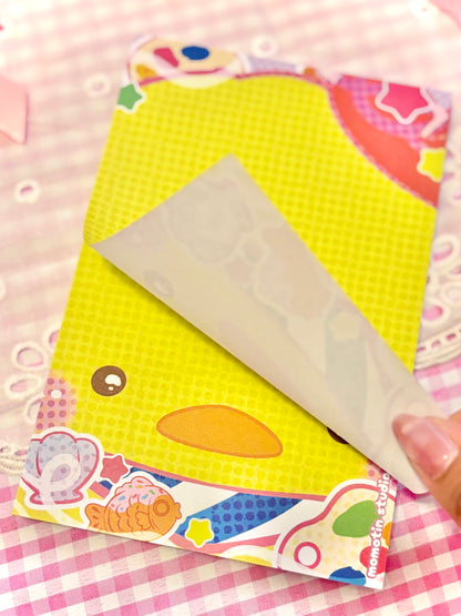 A close-up of a yellow duck-themed notepad with a page being lifted by a hand, set on a pink and white checkered fabric background.