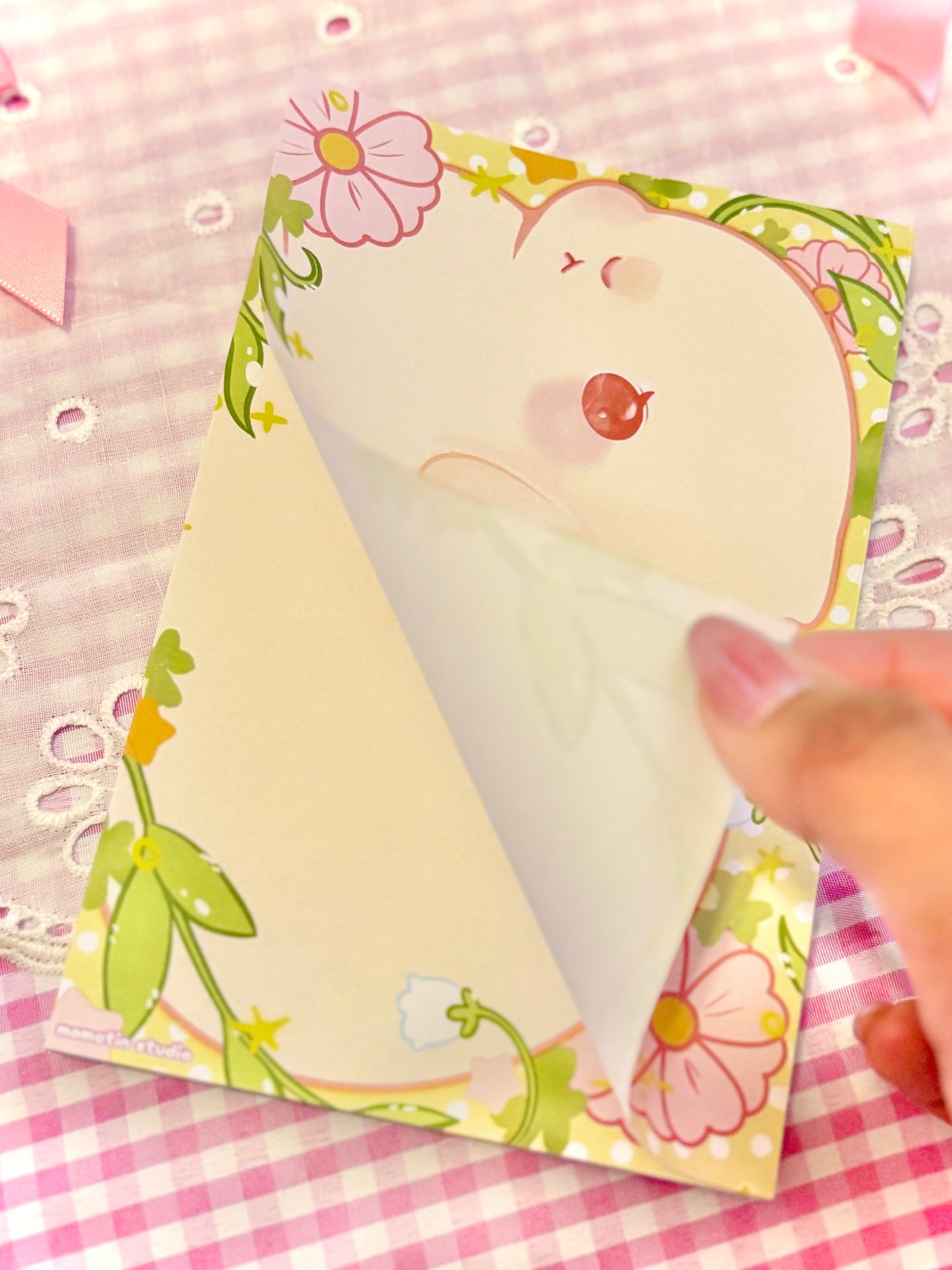 A hand peeling back a paper top layer of a pastel notepad featuring a cute white bunny with red eyes, set on a pink gingham fabric background.