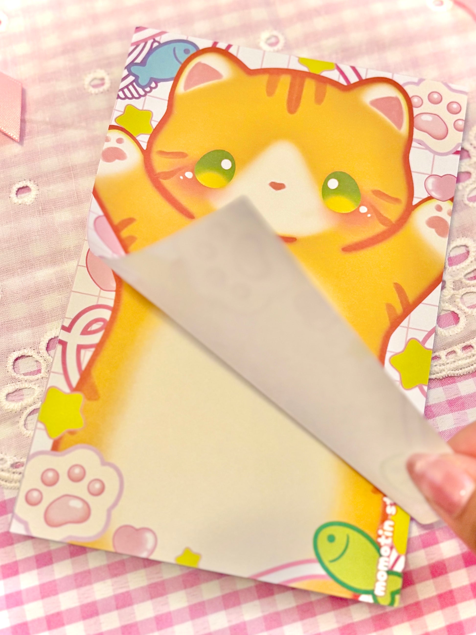 A close-up of a cute stationery notepad featuring a cartoon orange cat illustration, with a hand peeling back a translucent protective cover. The notepad is placed on a pink and white checkered fabric background.