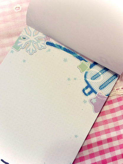 A close-up of a white notepad page with blue and pastel decorative graphics, partially curled at the top, resting on a pink and white gingham fabric surface.