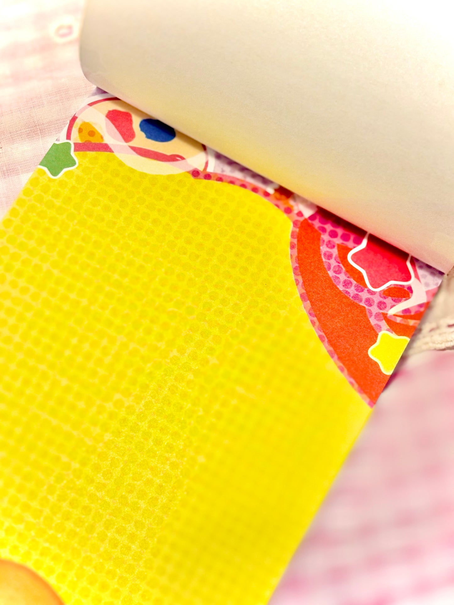 A close-up of a bright yellow notepad sheet partially unrolled, with a colorful patterned edge visible against a pink check fabric background.