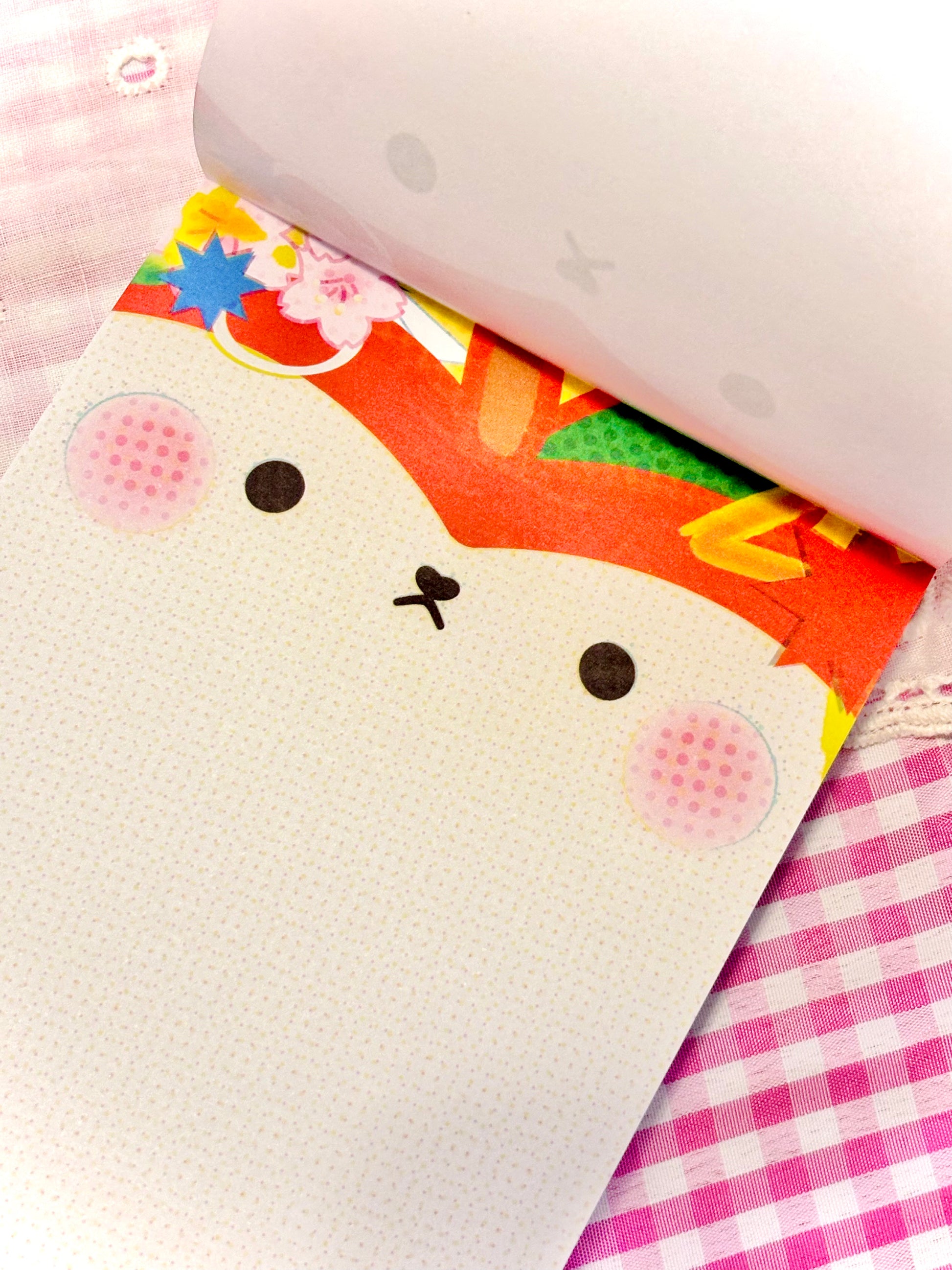 A close-up of a white textured notebook or journal with a cute cartoon apple face on the cover, partially open to reveal a colorful patterned interior, set on a pink and white checkered fabric surface.