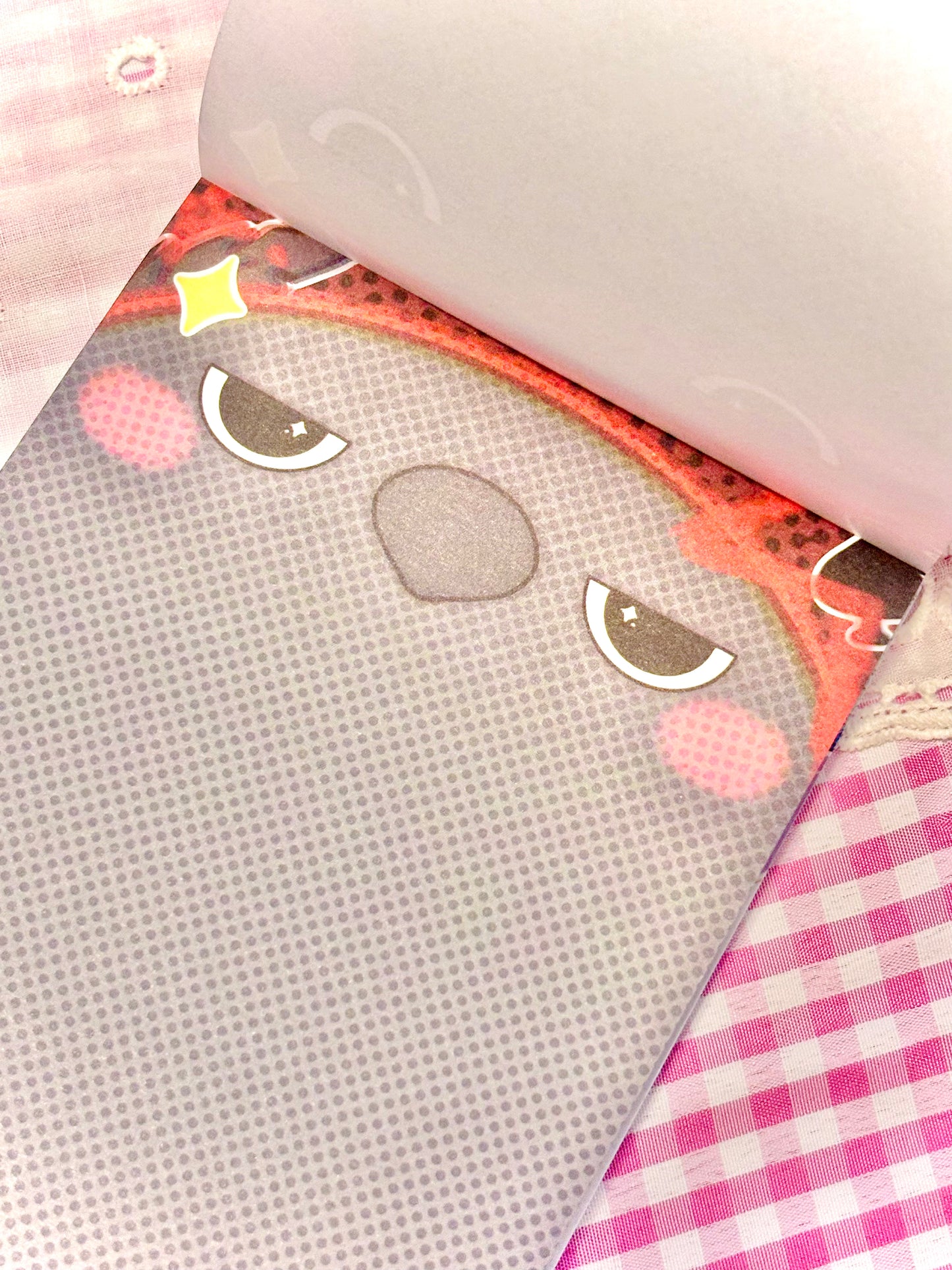 A close-up of a pink and white gingham fabric surface with a cute crow face notepad. The face has gray eyes with white outlines, a gray oval beak, pink cheeks, and a coral-red outline area with a small yellow star accent.