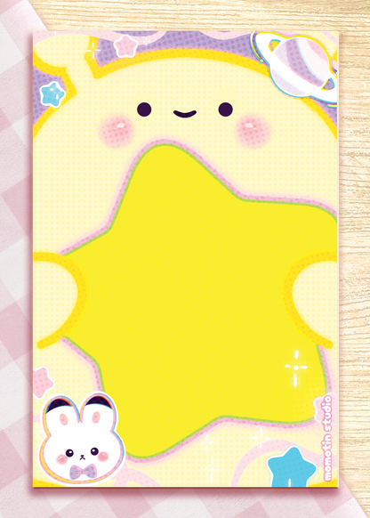 A cute pastel stationery notepad featuring a smiling yellow star-like character, pink cheeks, a small bunny at the bottom left, and a planet illustration at the top right, shown on a pink-and-white checkered surface over a light wood background.