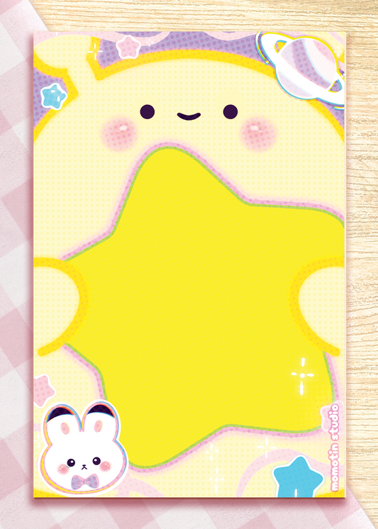 A cute pastel stationery notepad featuring a smiling yellow star-like character, pink cheeks, a small bunny at the bottom left, and a planet illustration at the top right, shown on a pink-and-white checkered surface over a light wood background.