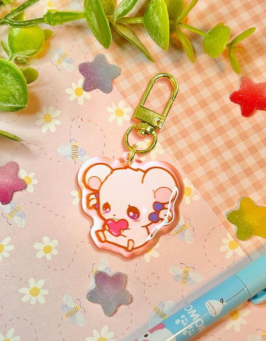A cute acrylic keychain charm shaped like a small white hamster holding a red heart, attached to a gold-tone lobster clasp. The charm is photographed on a pastel pink patterned surface with decorative flowers, leaves, and a blue pen nearby.