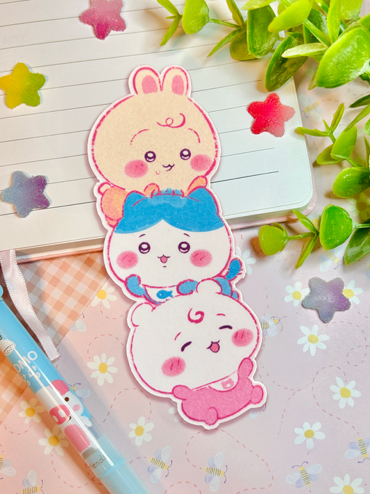 Three cute stacked bunny bookmarks with pink, blue, and white designs, arranged on a lined notebook with decorative green leaves and pastel star stickers.
