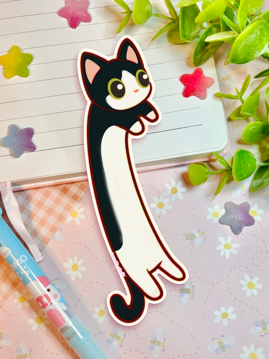 A die-cut sticker of a cute black and white cat with large eyes, shown as a flat-lay on a pink floral notebook with a pen, plant leaves, and star stickers.
