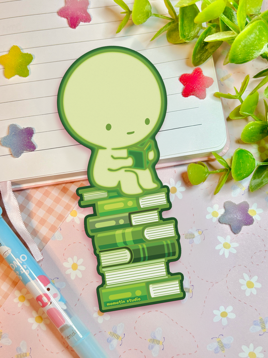 A green cartoon bookmark shaped like a round-headed character sitting on a stack of books, placed on a pink floral notebook with a pen, star stickers, and green leaves around it.