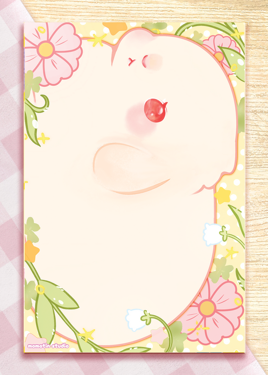 A pastel notepad featuring a cute white bunny with red eyes, surrounded by pink flowers and green leaves on a yellow border. The card is photographed on a pink-and-white checkered surface over a light wood background.