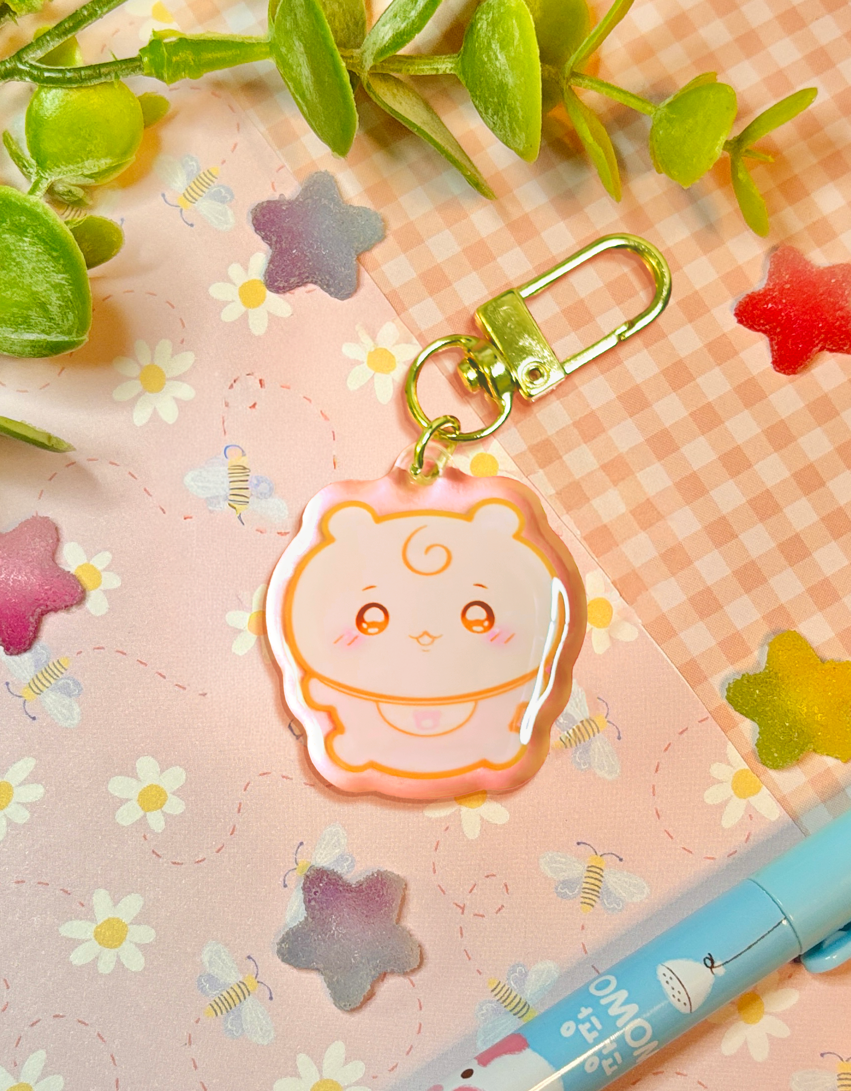 A cute pastel pink acrylic keychain charm with a white bear-shaped smiling cartoon character, attached to a gold metal key ring, displayed on a pink floral and gingham background with decorative leaves and a pen.