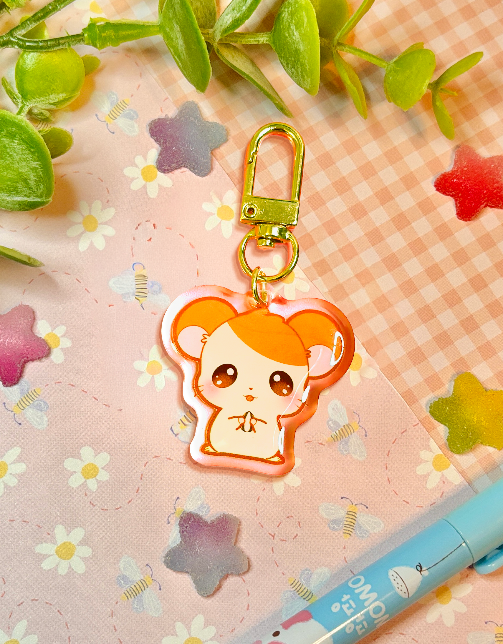 A cute cartoon hamster keychain charm with a gold clasp, resting on a pink floral and gingham background with decorative green leaves and a blue pen.