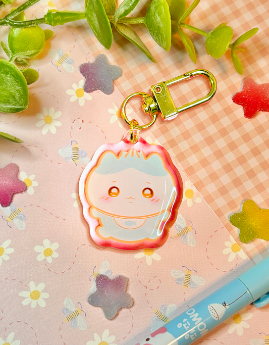 A cute pastel acrylic keychain charm featuring a smiling white and blue cat-like baby cartoon character with a gold clasp, displayed on a pink floral background with decorative leaves and a pen.