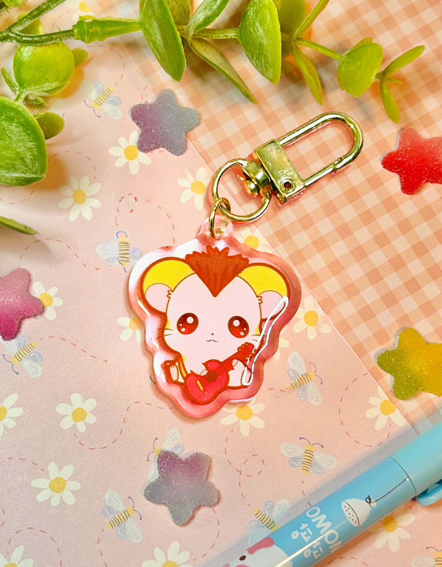 A cute anime keychain featuring a small yellow hamster with brown mohawk and a red guitar, attached to a gold clasp, displayed on a pink floral background with decorative leaves and a pen.