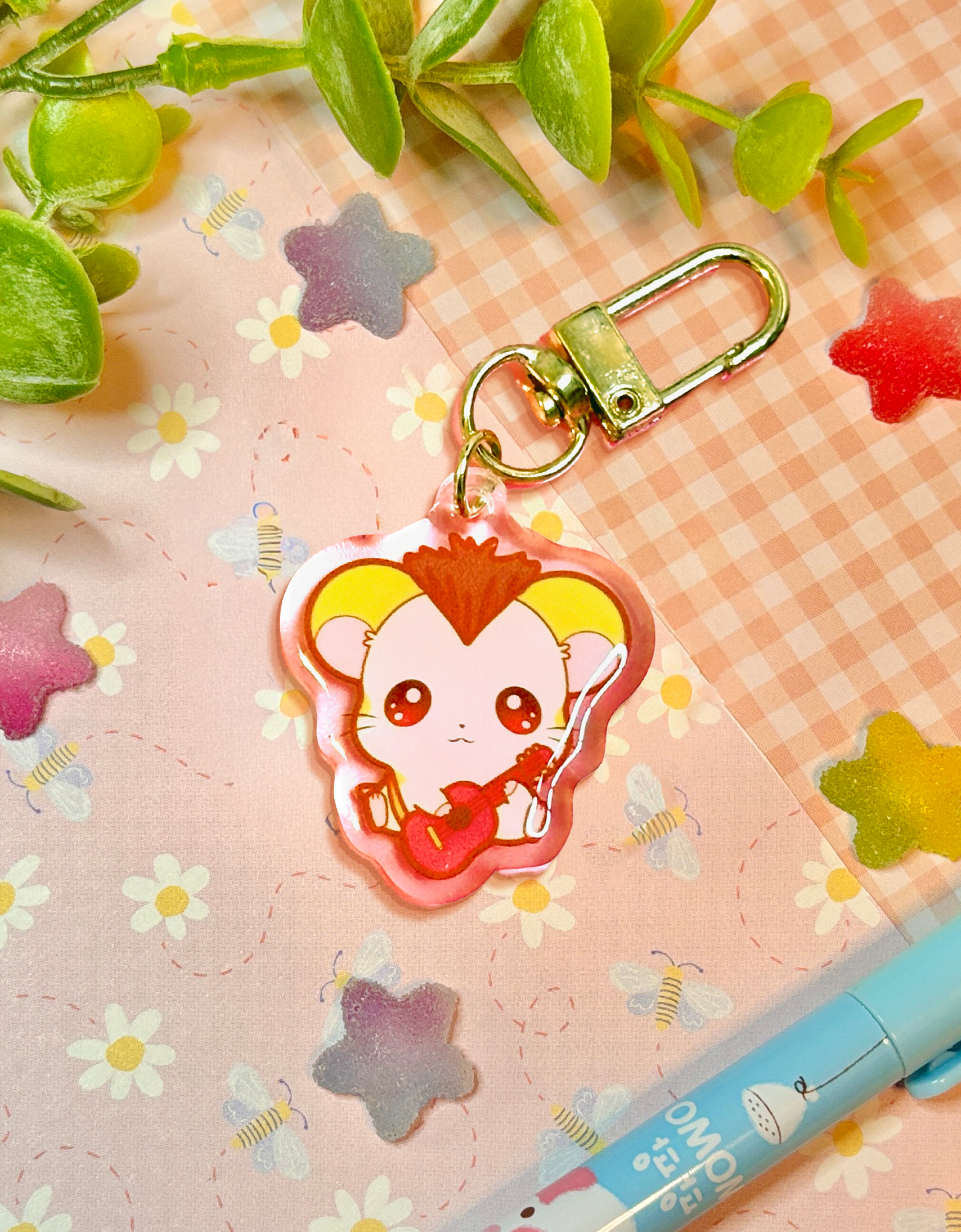A cute anime keychain featuring a small yellow hamster with brown mohawk and a red guitar, attached to a gold clasp, displayed on a pink floral background with decorative leaves and a pen.