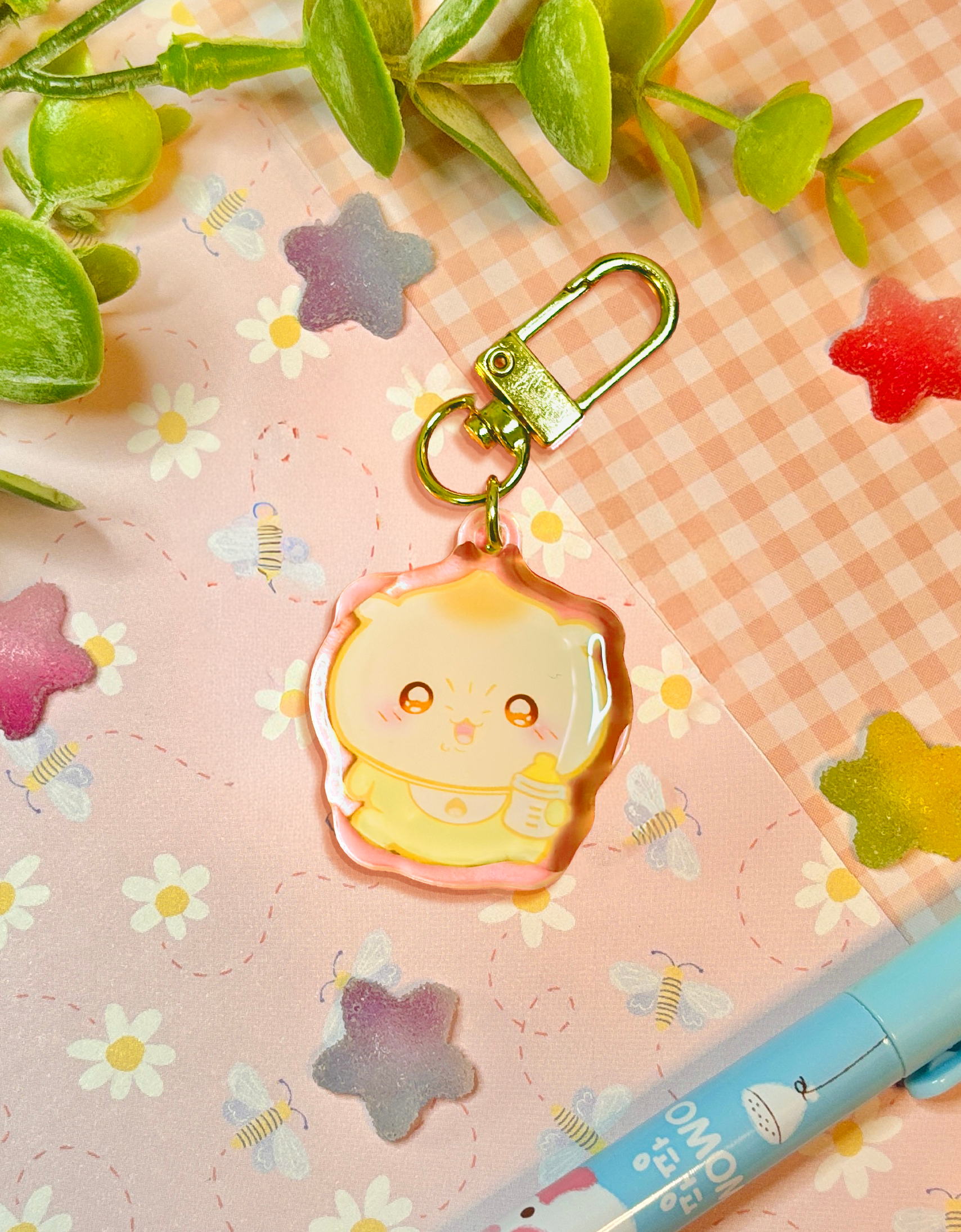 A cute pastel acrylic keychain featuring a scowling baby character with a yellow onesie and baby bottle, attached to a gold metal clasp. The keychain is displayed on a pink gingham paper background decorated with small white daisies, pastel star stickers, and green leaves.
