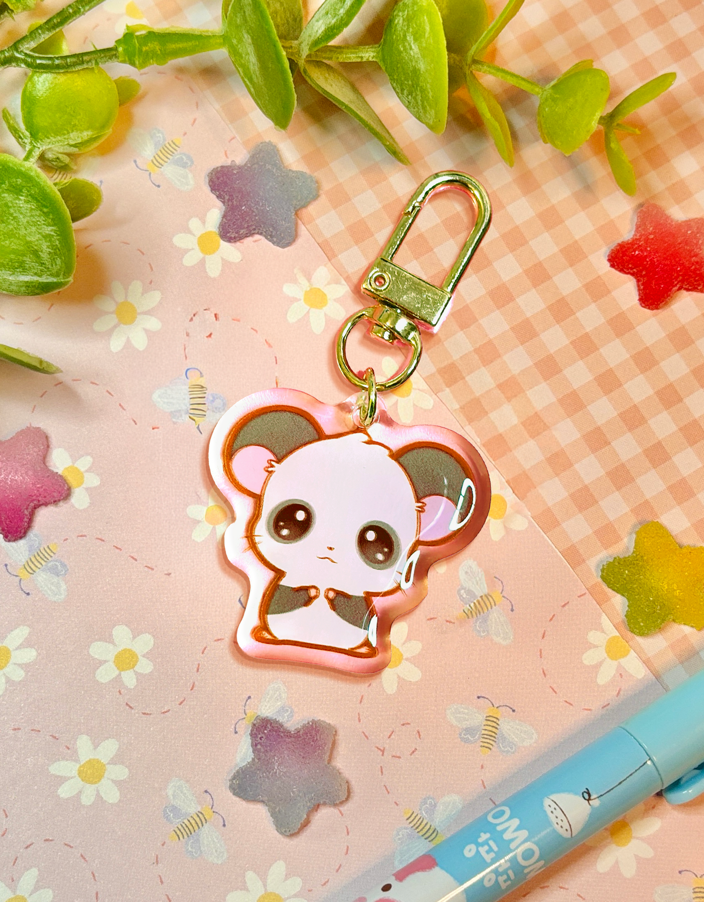 A cute cartoon panda keychain charm with a gold clasp, displayed on a pink floral notebook with decorative green leaves and a blue pen.