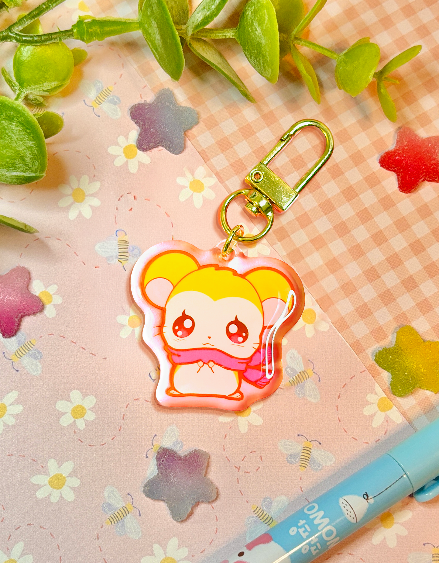 A cute anime keychain featuring a yellow-haired hamster with a pink scarf, attached to a gold metal clasp, displayed on a pink floral background with decorative green leaves and a blue pen.