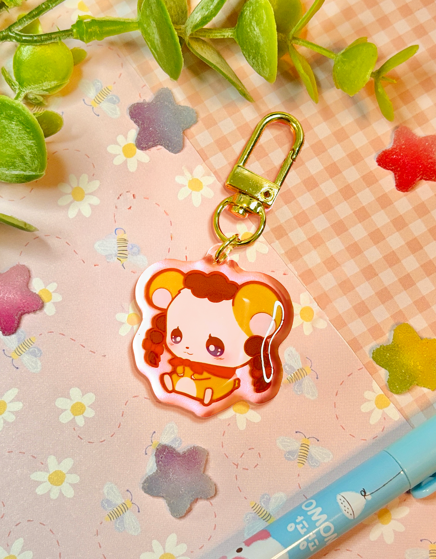 A cute cartoon keychain charm featuring a chibi hamster with brown puffy hair and a red scarf, attached to a gold metal clasp. The charm is displayed on a pink floral and gingham-patterned background with decorative green leaves and a blue pen.