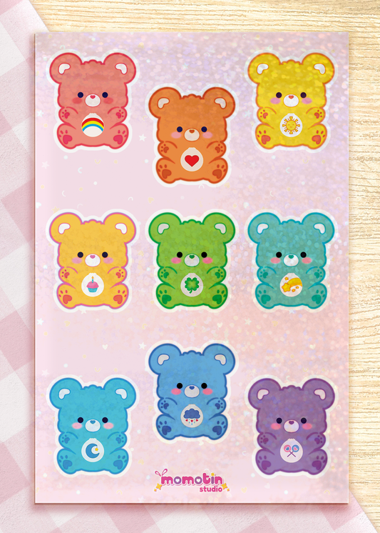 A sheet of nine cute Rainbow Bear stickers on a sparkly pink backing laminate, displayed on a light wood surface. The bears are in various pastel colors (pink, orange, yellow, green, teal, blue, purple) and each has a different symbol on its belly.
