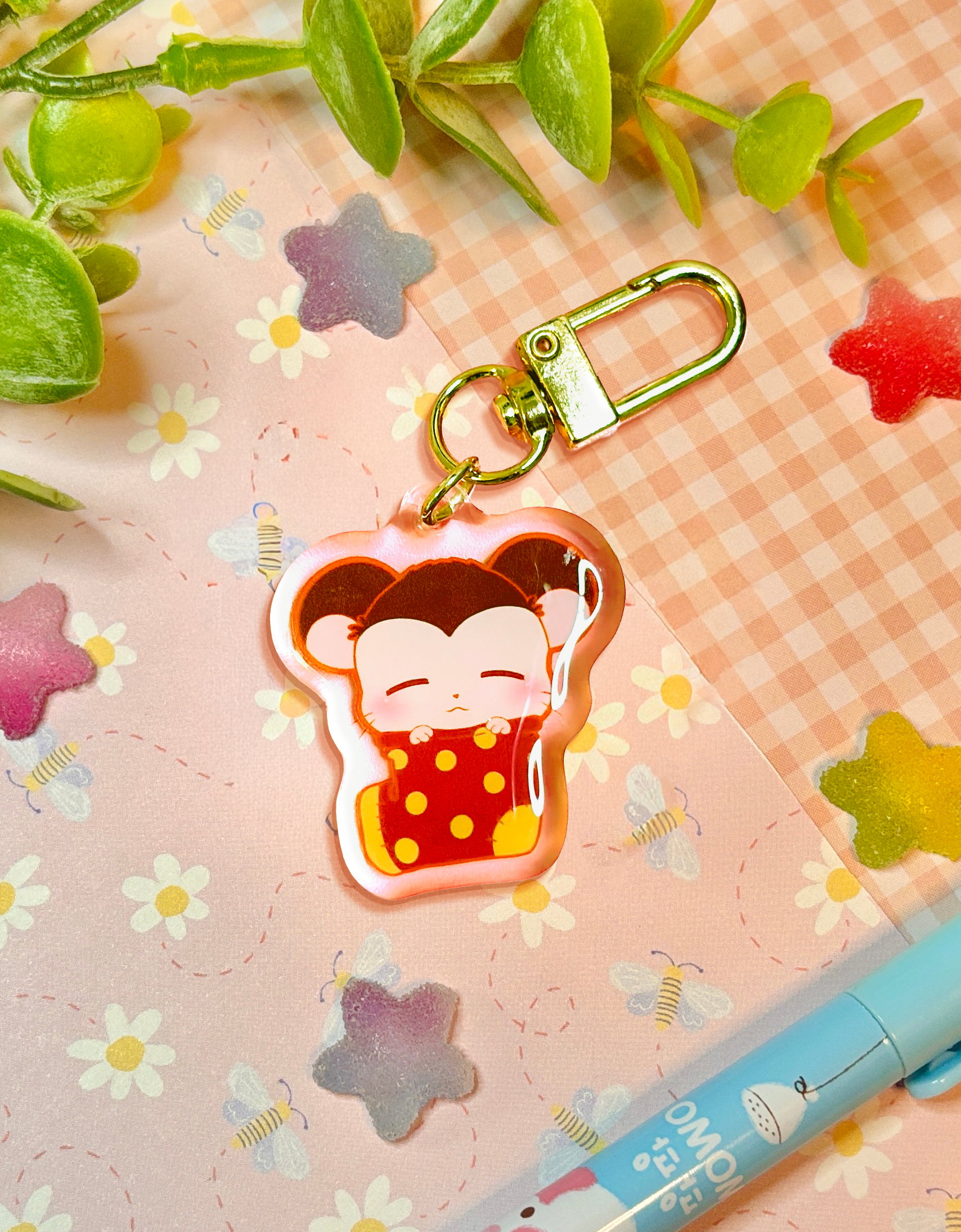 A cute cartoon hamster sleeping in an oversized red sock with yellow dots acrylic keychain charm with a gold clasp, displayed on a pink gingham paper background decorated with small flowers, leaves, and a blue pen.