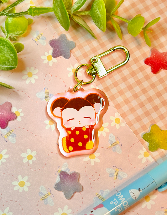A cute cartoon hamster sleeping in an oversized red sock with yellow dots acrylic keychain charm with a gold clasp, displayed on a pink gingham paper background decorated with small flowers, leaves, and a blue pen.