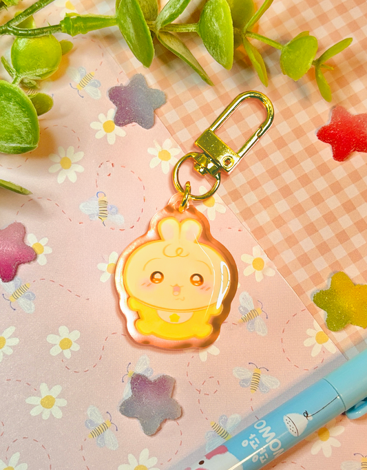A cute yellow cartoon baby bunny character acrylic keychain with a gold clasp, resting on a pink floral and gingham background with decorative green leaves and a blue pen.